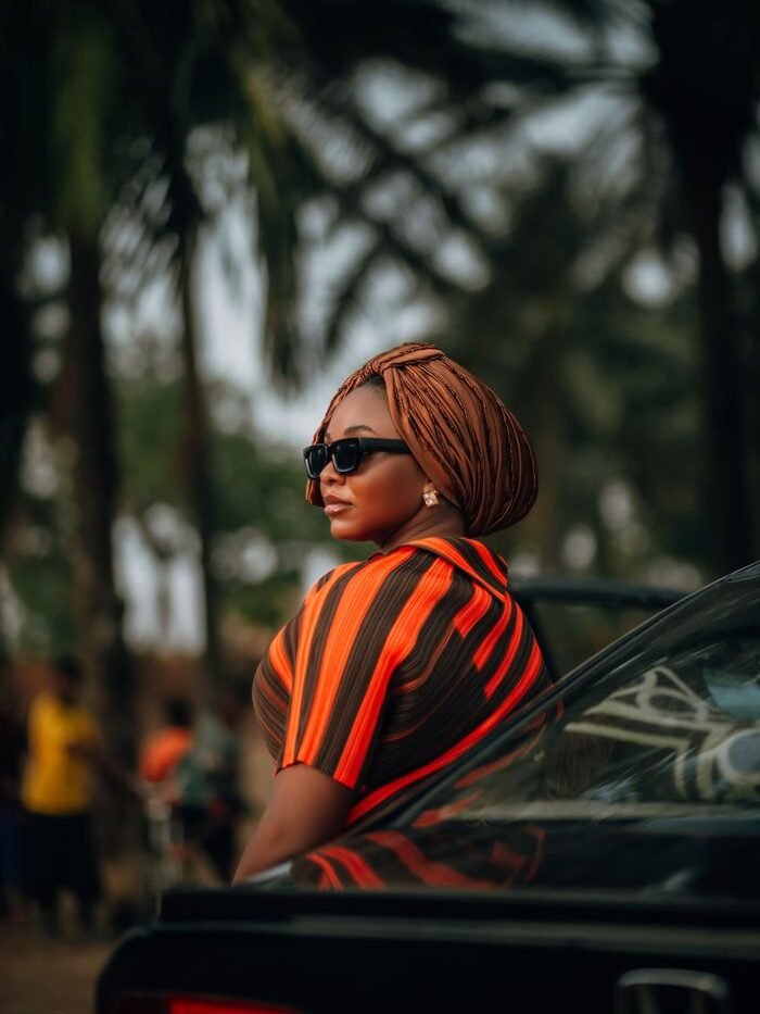 Confident woman wearing sunglasses and headwrap, standing by a car in a tropical outdoor setting.