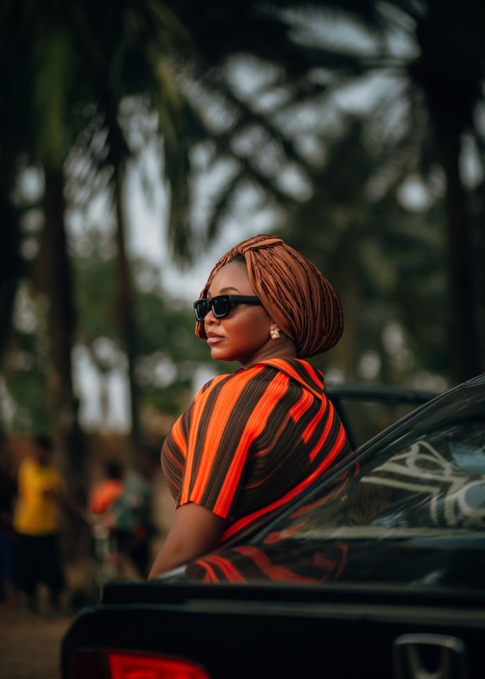 Confident woman wearing sunglasses and headwrap, standing by a car in a tropical outdoor setting.